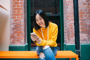 Genç, kısa saçlı, sarı kapüşonlu ve yırtık pantolonlu bir kadın. Telefondan mesaj atıyor. Bankta oturuyor ve ekrana bakıyor.