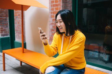 Genç, kısa saçlı, sarı kapüşonlu ve yırtık pantolonlu bir kadın. Telefondan mesaj atıyor, bankta oturuyor, gülümsüyor ve ekrana bakıyor.