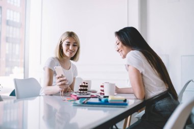 Kadın arkadaşların cep telefonlarını paylaşırken, masada beyaz ışıklı odada not defterleriyle toplanan çay ve keklerin keyfini çıkarırken yan görüntüsü