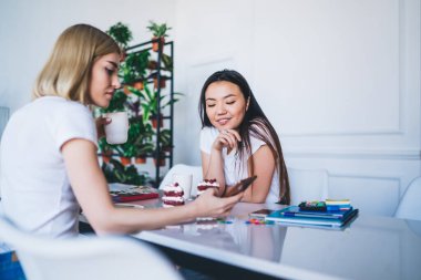 Beyaz tişörtlü çok ırklı kadınlar cep telefonlarıyla mesajlaşıp, ellerinde kahve ve pastalarla modern yaratıcı mekanda oturuyorlar.