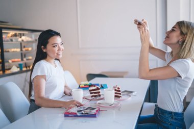 Masada oturan beyaz tişörtlü çeşitli kadınlar kafeteryada arka planda tezgahı olan kahve bardakları ve not defterleriyle akıllı telefonlarıyla fotoğraf çekiyorlar.