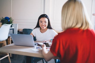 Dizüstü bilgisayarla çalışan, cep telefonuyla sörf yapan, modern ışık yaratıcı çalışma ortamında masada oturan çok ırklı gülümseyen neşeli kadınlar. 