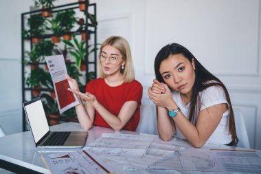 Gözlüklü genç sarışın iş yerindeki kameraya bakarak günlük kıyafetlerle etnik kadınlarla işbirliği projesi yapıyor. 