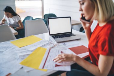 Sıradan kıyafetli bir kadın ofiste boş ekranlı dizüstü bilgisayarla masada otururken akıllı telefon konuşmaları yapıyor ve belgeleri inceliyor.