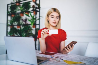 Akıllı, düşünceli bir kadın elinde kartvizitle çalışıyor ve ofiste cep telefonuyla sörf yapıyor. 
