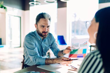 Sıradan giyinen bir erkek, çalışma alanında birlikte çalışırken masada oturup kadın meslektaşına diyagramı açıklar.