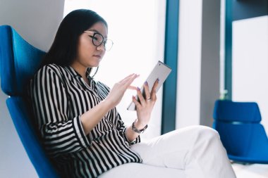 Resmi çizgili bluz ve beyaz pantolonlu, ofis koltuğunda oturan ve tablet üzerinde internette gezinen çekici genç bayanın yan görüntüsü