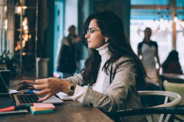 Uzun siyah saçlı, rahat giyimli, laptopla çalışan, etiketli ve kafede not defteri olan genç ve zeki bir bayan.  