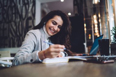 20 'li yaşlardaki neşeli beyaz kadın eğitim projesini incelerken gülümsüyor, modern uzayda bilgisayar ve ekipmanla bilgi öğrenmek ve öğrenmek için oturan mutlu hipster öğrenci.