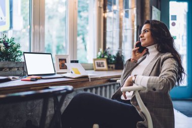 Genç, zeki, neşeli, uzun siyah saçlı, sıradan giyinmiş, cep telefonuyla konuşan, dizüstü bilgisayar işaretleriyle masada oturan mutlu bir bayan.