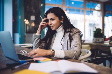 Koyu renk uzun saçlı, ceketli ciddi bir kadına yoğunlaşın. Laptop 'a göz gezdirin. Kahve dükkanında not defteri çıkartmalarıyla birlikte masada oturun.