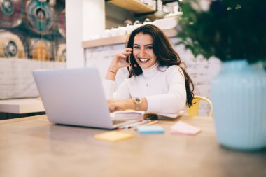 Mutlu, uzun siyah saçlı, beyaz kazaklı, cep telefonuyla konuşan, bilgisayar etiketli, yaratıcı iş yerinde kalemleri olan mutlu bir bayan. 