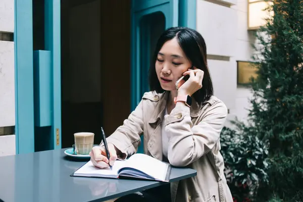 专注的年轻女性 黑发短发 坐在办公桌前 一边喝咖啡 一边用智能手机在笔记本上写字 — 图库照片