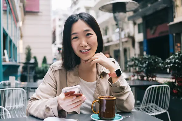 Wesoła Azjatka Luźnych Ubraniach Przeglądająca Smartfona Siedząc Przy Stole Filiżanką — Zdjęcie stockowe