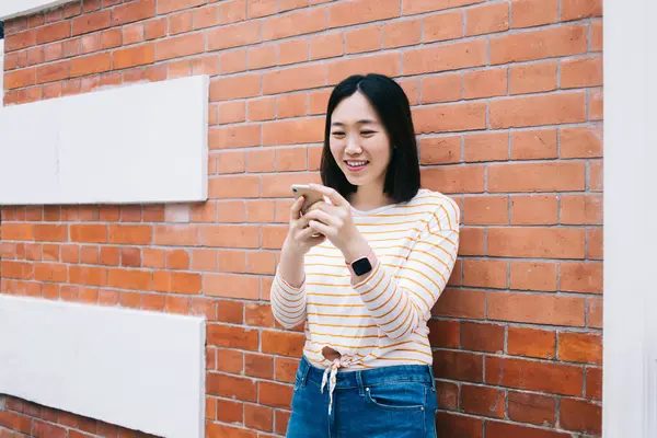 Keyifli genç etnik kadın, günlük kıyafetleriyle tuğla bina duvarına yaslanıyor ve şehir sokaklarında modern akıllı telefon kullanıyor.
