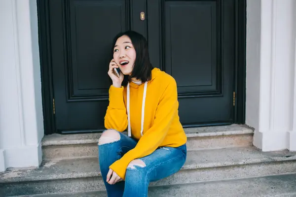 Amused Asian female in yellow hoodie and denim resting on street steps while chatting on mobile phone and smiling away