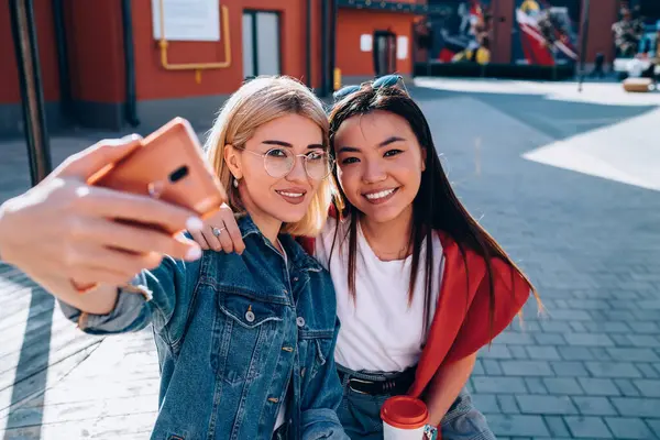 Gündelik giysiler içinde genç sırıtan kadınlar bir fincan kahveyle şehirde yaz güneşli bir günde akıllı telefonla selfie çekiyorlar.