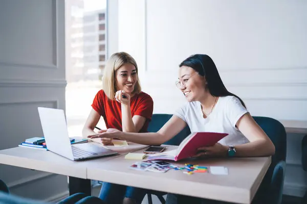 stock image Young smart smiling multiracial coworkers in casual apparel working with laptop sitting at table making notes in notepad  in modern white studio