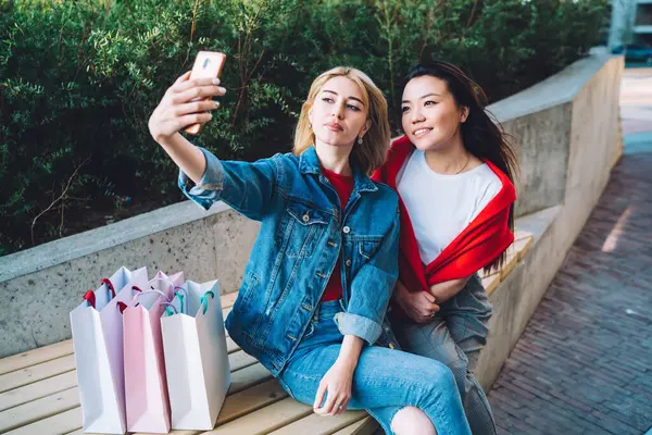 Yukarıdan gelen memnun edici, çok ırklı, gündelik kıyafetlerle çekici kız arkadaşlar akıllı telefondan selfie çekerken caddede bankta yan yana oturuyorlar.