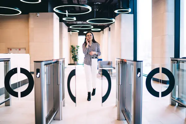 Young confident woman in striped blouse and white pants talking on smartphone while leaving modern office center through automatic turnstile gates