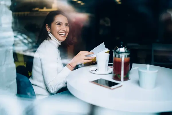 Cheerful female reader holding literature poetry book in hands and laughing during leisure time for recreation with novel, happy hipster girl smiling from positive plot sitting at cafeteria table