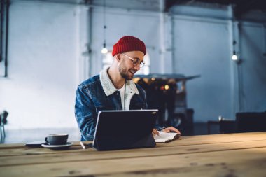 Kırmızı şapkalı, kot ceketli, hırslı, zeki, gülümseyen yetişkin işçi modern ofisteki dizüstü bilgisayarın yanına, ahşap masaya oturmuş notlar yazıyor.