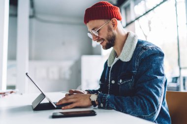Genç, şık, sakallı, gözlüklü ve şapka takan erkek öğrenci. Beyaz odadaki tablete mesaj yazıyor. Arka planda bulanıklık var.