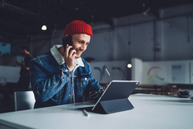 Genç, sakallı, serbest çalışan, sıcak elbiseli, şapkalı, iş yerinde telefonla konuşan, gülümseyen ve tablet ekrana bakan bir erkek.