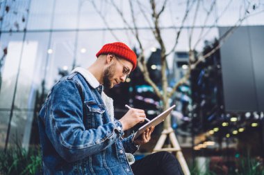 Caddede cam gökdelene karşı oturan ve Stylus 'la grafik tabletine proje çizen modaya uygun sakallı bir adam. 
