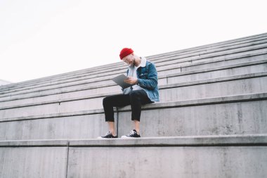 Gözlüklü, kırmızı şapkalı ve kot ceketli güler yüzlü, şık sakallı genç adam beton bankta oturuyor ve açık havada tablete vuruyor.