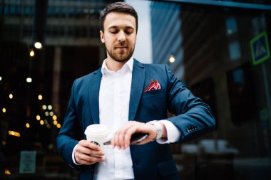 Şık mavi takım elbiseli, meşgul bir girişimci, modern şehir caddesinde kol saatini kahveyle kontrol ediyor. 