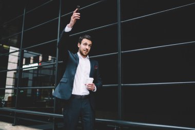 Resmi kıyafetli, elinde telefonla bir fincan kahve içen, modern şehrin sokaklarında el sallayan yakışıklı bir erkek.