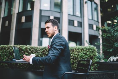 Resmi takım elbiseli akıllı çalışanın yan görünüşü dizüstü bilgisayarda çalışması ve şehir kafeteryasında akıllı telefonlara göz atması.