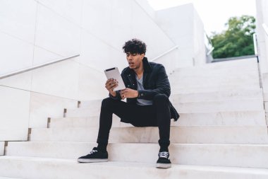 Modern stil ile ilgilenen Afrikalı Afrikalı Afrikalı Amerikalı bir adam gün ışığında şehir merdivenlerinde otururken tablet üzerinde geziniyor.