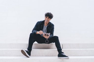 Fotokopi duvarının yanında dinlenen, kameraya gülümseyen dijital tabletli pozitif melodiyi seven neşeli Latin hippi adam portresi.