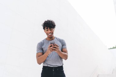 Gri tişörtlü, yakışıklı, kıvırcık, yetişkin bir erkek beyaz duvarın önünde sokakta akıllı telefonuna göz atıyor.  