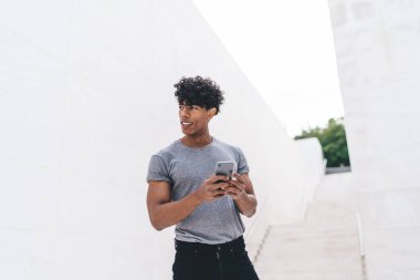 Gülümseyen, kıvırcık, gri tişörtlü genç adam, beyaz duvarın önündeki merdivenlerde cep telefonunu karıştırıyor. 