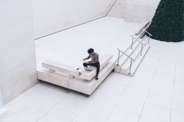 Beyaz iş merkezindeki dizüstü bilgisayarın önündeki bankta oturan gündelik kıyafetlerle belgelere bakan yukarıdaki etnik adamdan.