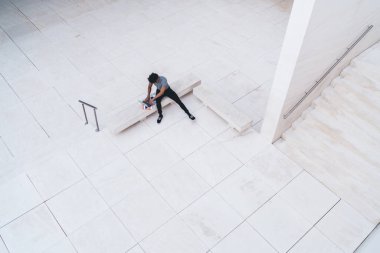 Üst kattaki erkek, günlük kıyafetlerini giyip dizüstü bilgisayarla caddedeki mermer bahçedeki bankta oturmuş belgeleri kontrol ediyor. 