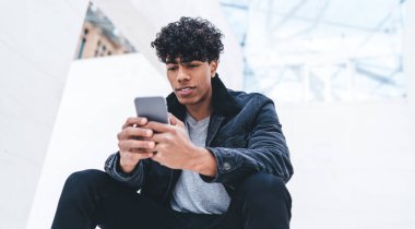 Tarz sahibi, kıvırcık saçlı, şık giyinmiş, beyaz, modern bir binada otururken akıllı telefon kullanan, modaya uygun, Afro-Amerikan bir adam.