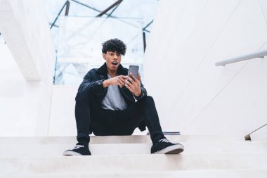 Beyaz modern binada merdivenlerde otururken kıvırcık saçlı afro-amerikalı genç adam akıllı telefon kullanıyor.