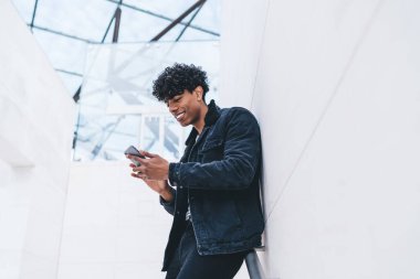 Sosyal medyada video izlerken ve gülümserken akıllı telefonuyla beyaz duvara yaslanan şık kotlu, neşeli, kıvırcık siyah adamdan.