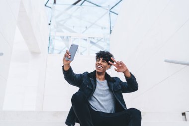 Akıllı telefonlu mutlu kıvırcık saçlı Amerikalı. Parlak odada selfie çekiyor ve V işareti gösteriyor.