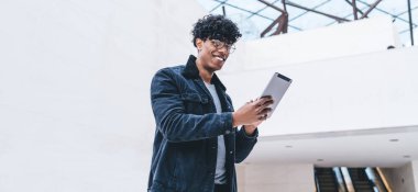 Kot takım elbiseli, tabletli gözlüklü neşeli, kıvırcık, siyah adam büyük bir odada kameraya bakıyor.