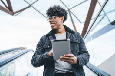 Siyah sıcak ceketli yakışıklı, kıvırcık erkek. Yuvarlak gözlüklü. Yürüyen merdivene tabletle doğru gidiyor. 