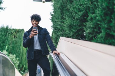 Siyah ceketli ve kot pantolonlu mutlu, kıvırcık, yetişkin bir erkeğin arka plandaki yeşil ağaçlarda asansörde akıllı telefon kullanmasından. 