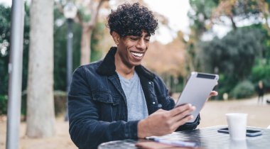 Keyifli etnik adam, günlük giysiler içinde, neşeyle tableti karıştırıyor, parktaki kafe masasında tek başına vakit geçirirken.