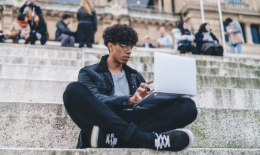 Düşünceli, gözlüklü, sıradan kıyafetler giyen, özenle dizüstü bilgisayar kullanan, arka planda insanlarla birlikte şehir caddesinde oturan etnik erkek.