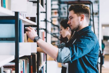 Ciddi sakallı, kot gömlekli, kol saati takan şehir kütüphanesinde kitap seçen ciddi bir gencin yan görüntüsü. 