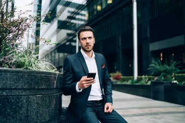 Thoughtful man in elegant suit using phone messaging with partner surfing social media relaxing from work sitting in business district looking away